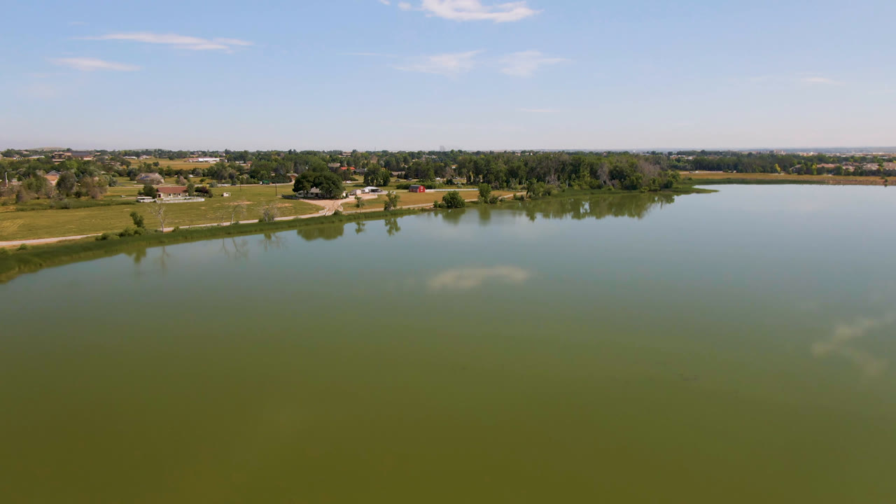 Aerial view of lake water leading to grassy shoreline, trees, houses, and distant land.