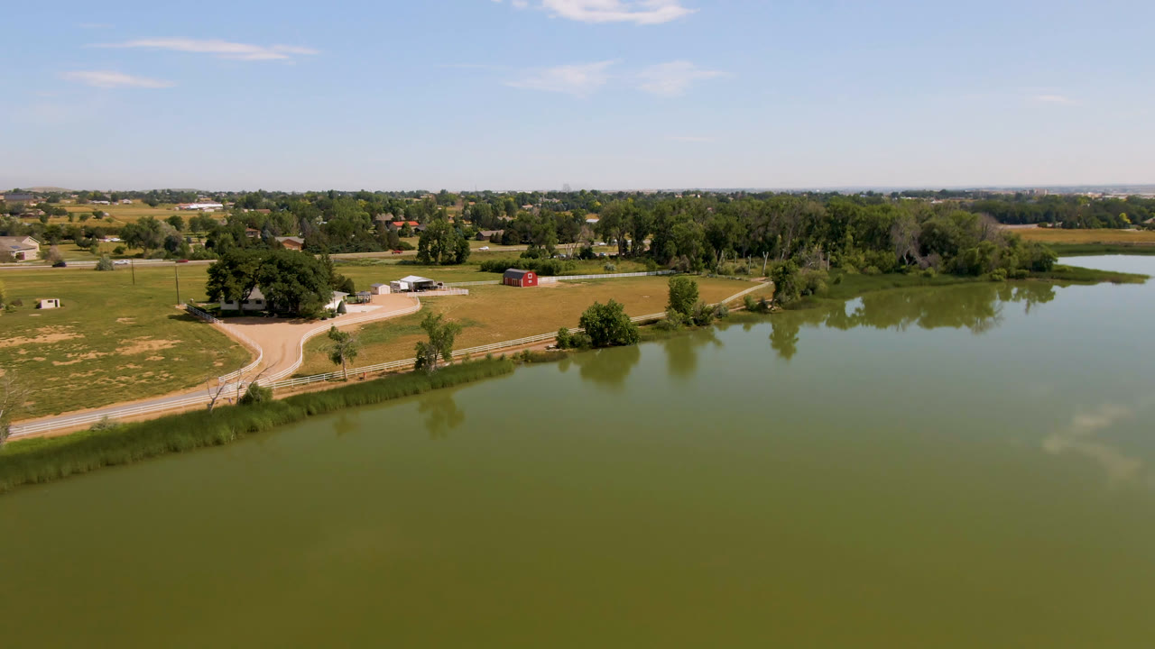Broomfield, Colorado drone view with the water covering more of the lower frame, a curving rural road at left, tree clusters near small buildings, and no visible people.
