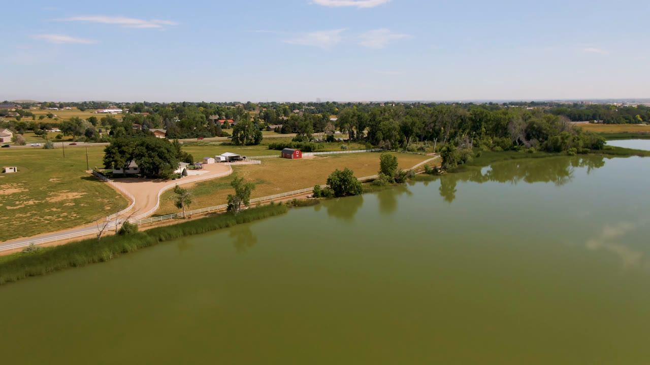 Wide aerial of green water beside fields, a roadside fence, trees, and distant buildings.