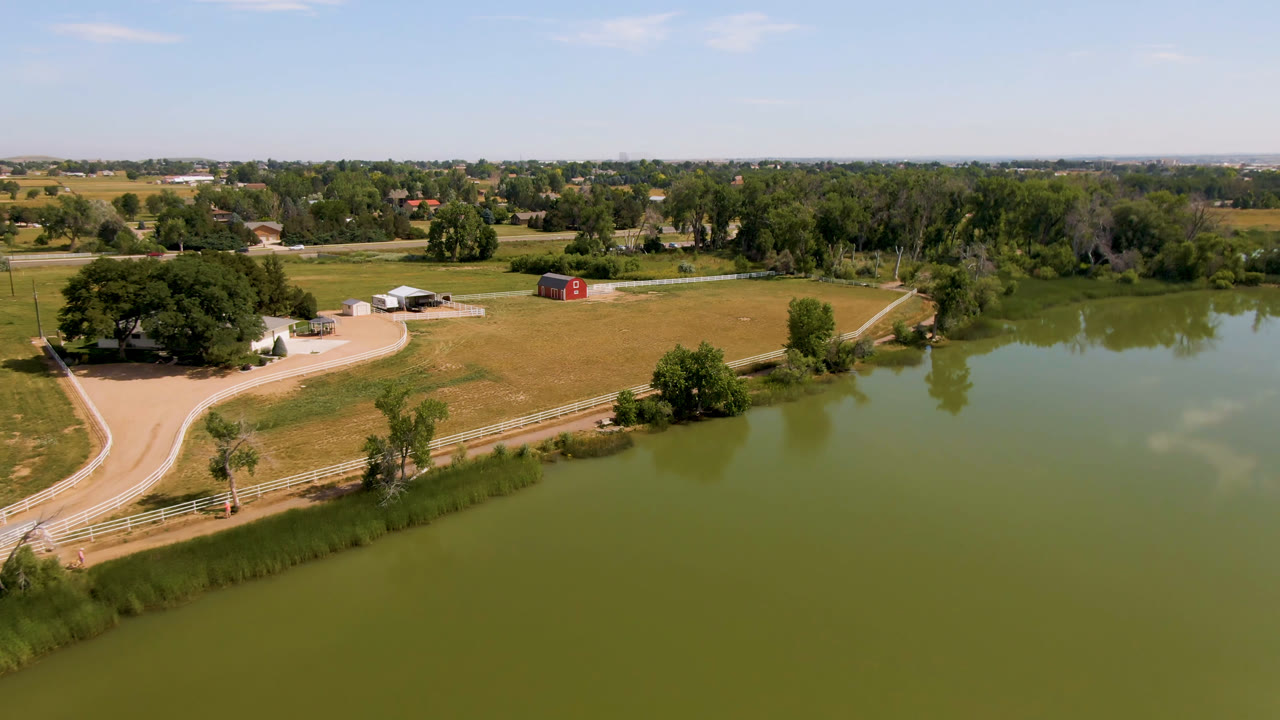 Aerial view in Broomfield, Colorado showing green pond water, fenced pasture, a curving road, red and white farm buildings, trees, and no visible people at the start of the clip.