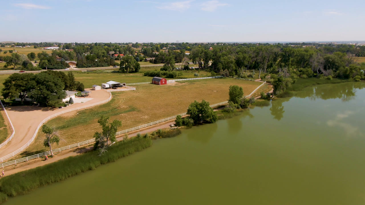 Drone-wide frame in Broomfield, Colorado with the pond stretching across the foreground, tan pasture and fence lines beyond it, a red barn near the center, and no visible people.