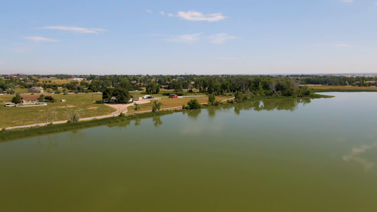 Early aerial frame over a green lake in Broomfield, Colorado, with a grassy shoreline, scattered trees, a curving road, distant buildings, blue sky, and no visible people.