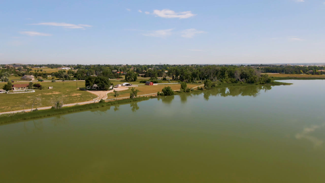 Second aerial frame shows more of the lake surface across the foreground in Broomfield, Colorado, with field edges, trees, small structures, and no visible people.