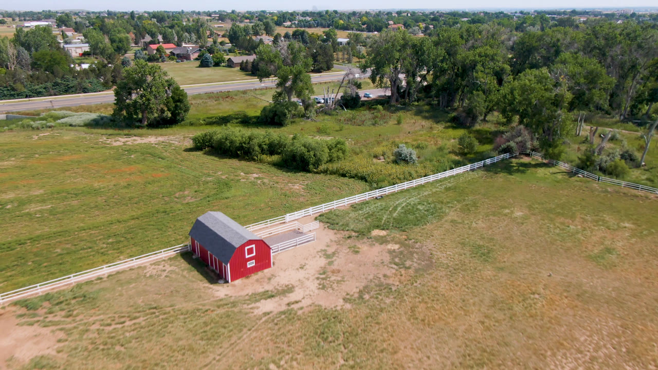Aerial frame from Broomfield, Colorado with the red barn lower left, white rail fencing crossing the field, a road in the upper frame, dense trees, distant houses, and no visible people.