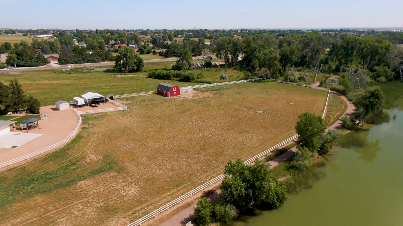 Aerial frame from Broomfield, Colorado showing a red barn left of center, dry fenced fields, green pond water on the right, distant roads, trees, homes, and no visible people.