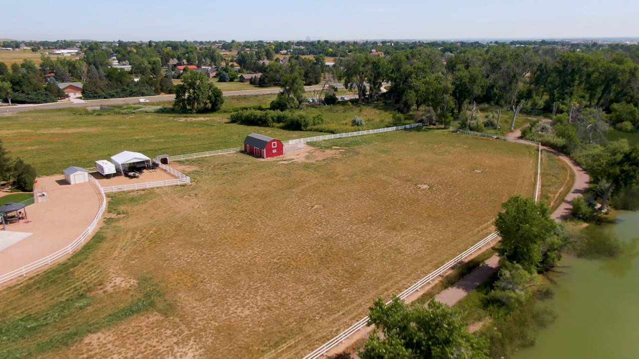Aerial view in Broomfield, Colorado with the red barn near the center-left, fenced paddock and outbuildings to the left, pond edge at right, dry grass field, distant tree line, and no visible people.