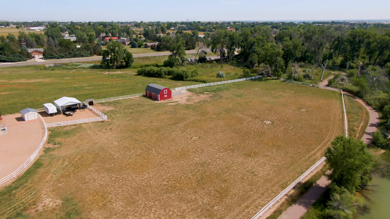 High aerial view over dry pasture with a red barn, pond, fence line, and roads beyond.