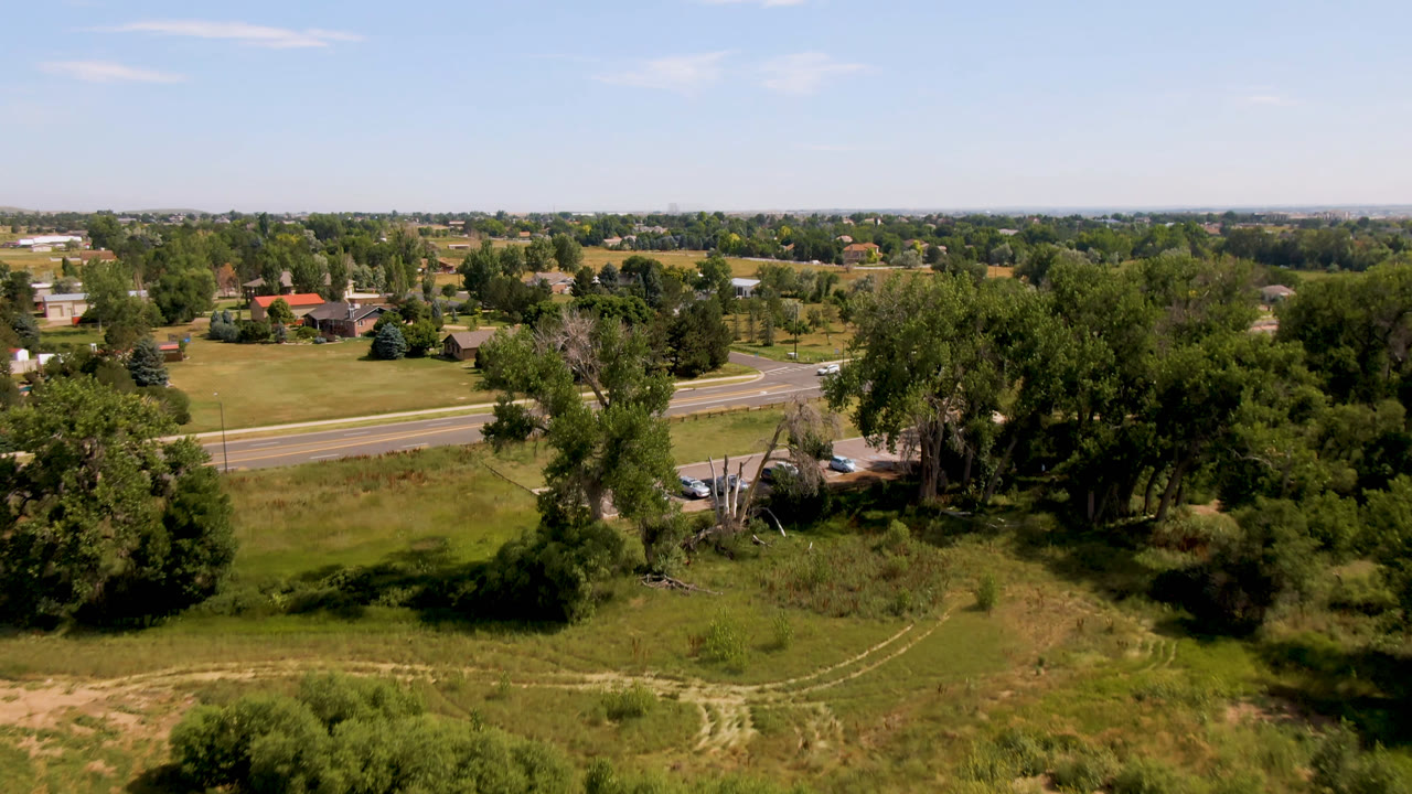 Wide aerial view in Broomfield, Colorado showing roadside trees, grassy open land, a divided road with small cars, distant homes, and no visible people.