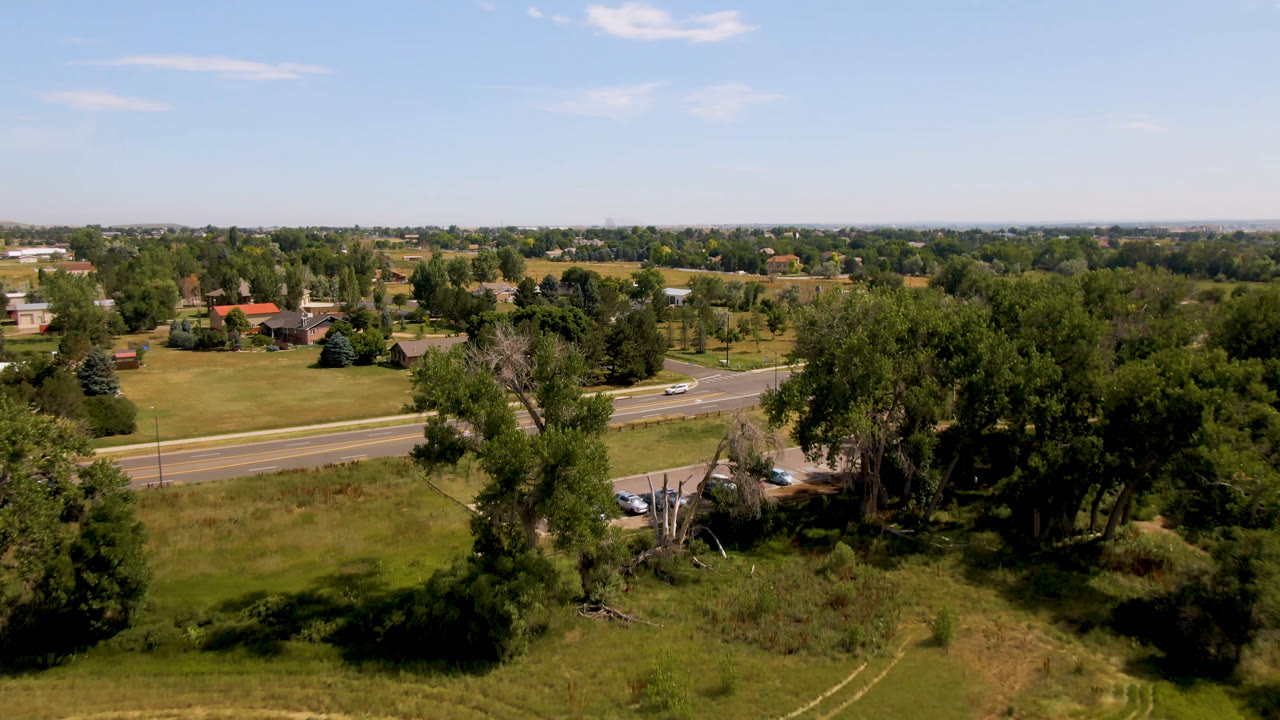 Broomfield, Colorado aerial frame with dense trees in the foreground, a divided road crossing the midground, lawns and houses beyond, and no visible people.