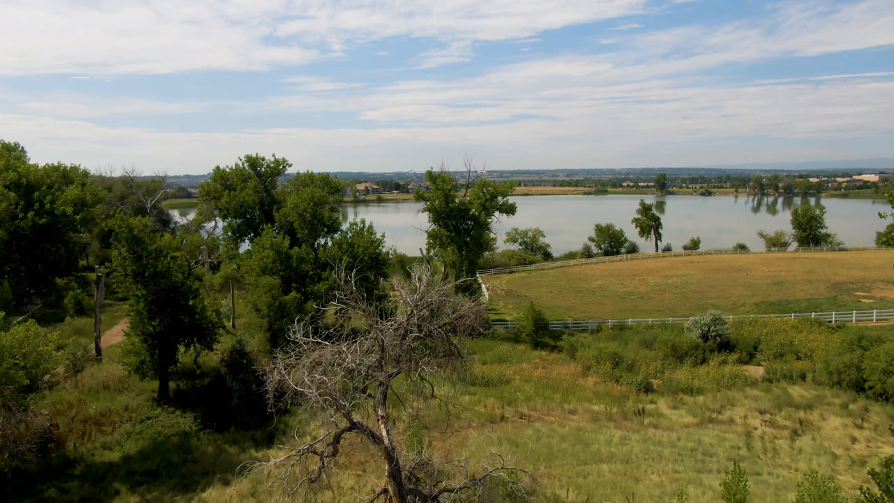 Elevated Broomfield, Colorado landscape with foreground trees, dry grass, a lake behind a field, distant low buildings, clouds, and no visible people.