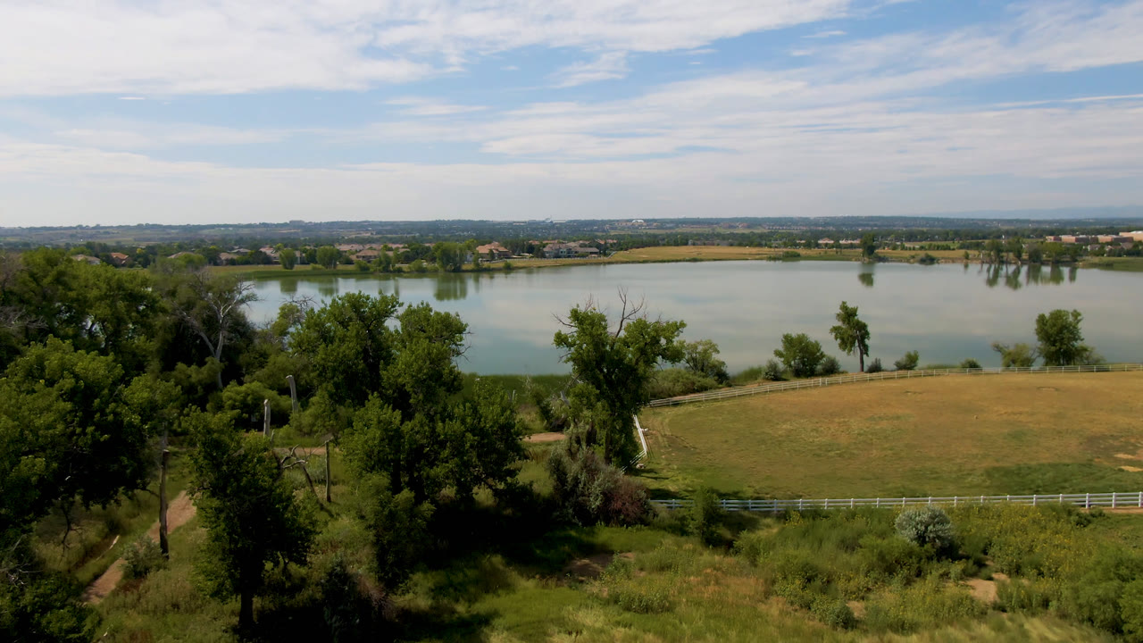 Elevated Broomfield, Colorado view with green trees in the foreground, a dry grassy field and white fence below, a pale lake beyond, and no visible people.
