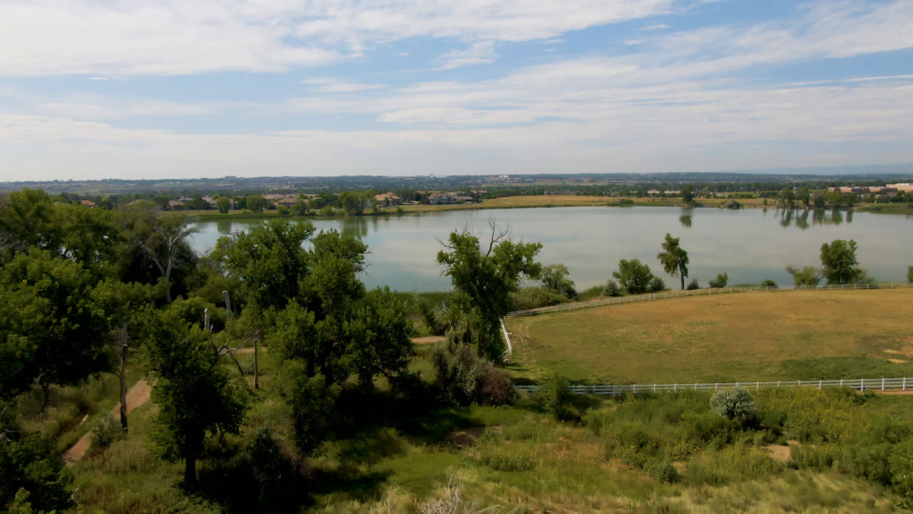 Wide Broomfield, Colorado landscape showing a lake across the background, scattered trees over a grassy slope, a white fence line, and no visible people.