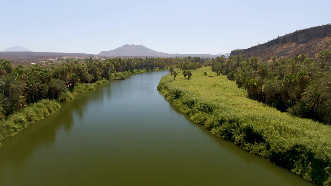 Wide aerial frame from San Ignacio with the river centered between bright grasses and tree cover, backed by a dry hillside, distant mountain, and no visible people.
