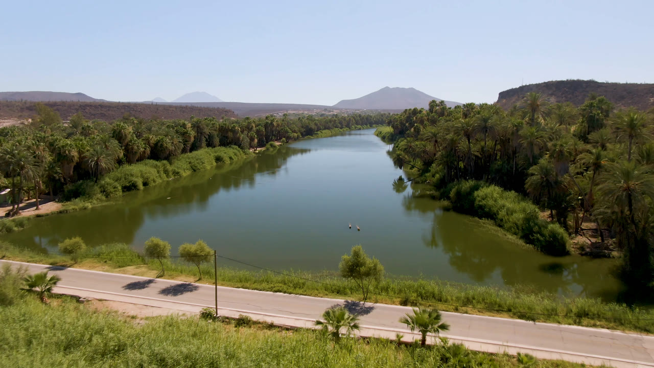 Wide aerial view of a green river, palms, roadside, and desert hills.