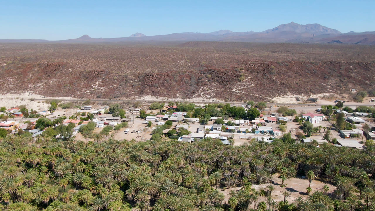 Wide aerial view of palms, low buildings, desert ridge, and distant mountains.