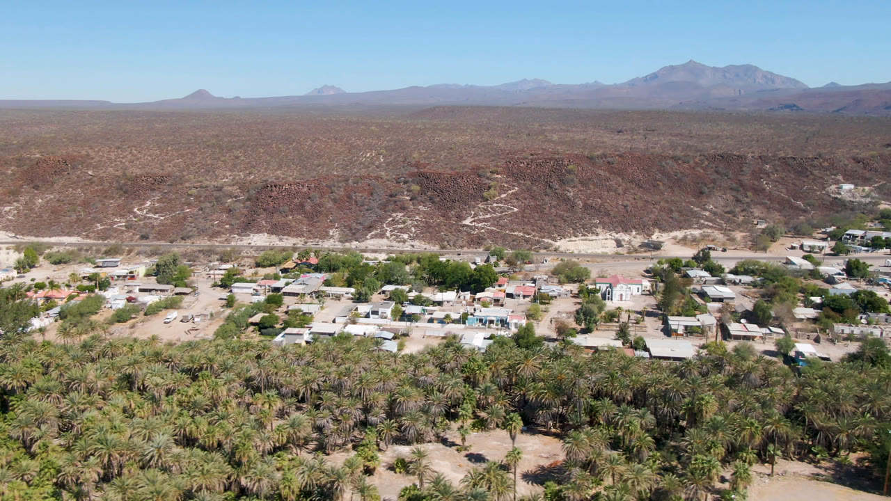 Aerial view of palm trees, small town buildings, desert ridges, and distant mountains