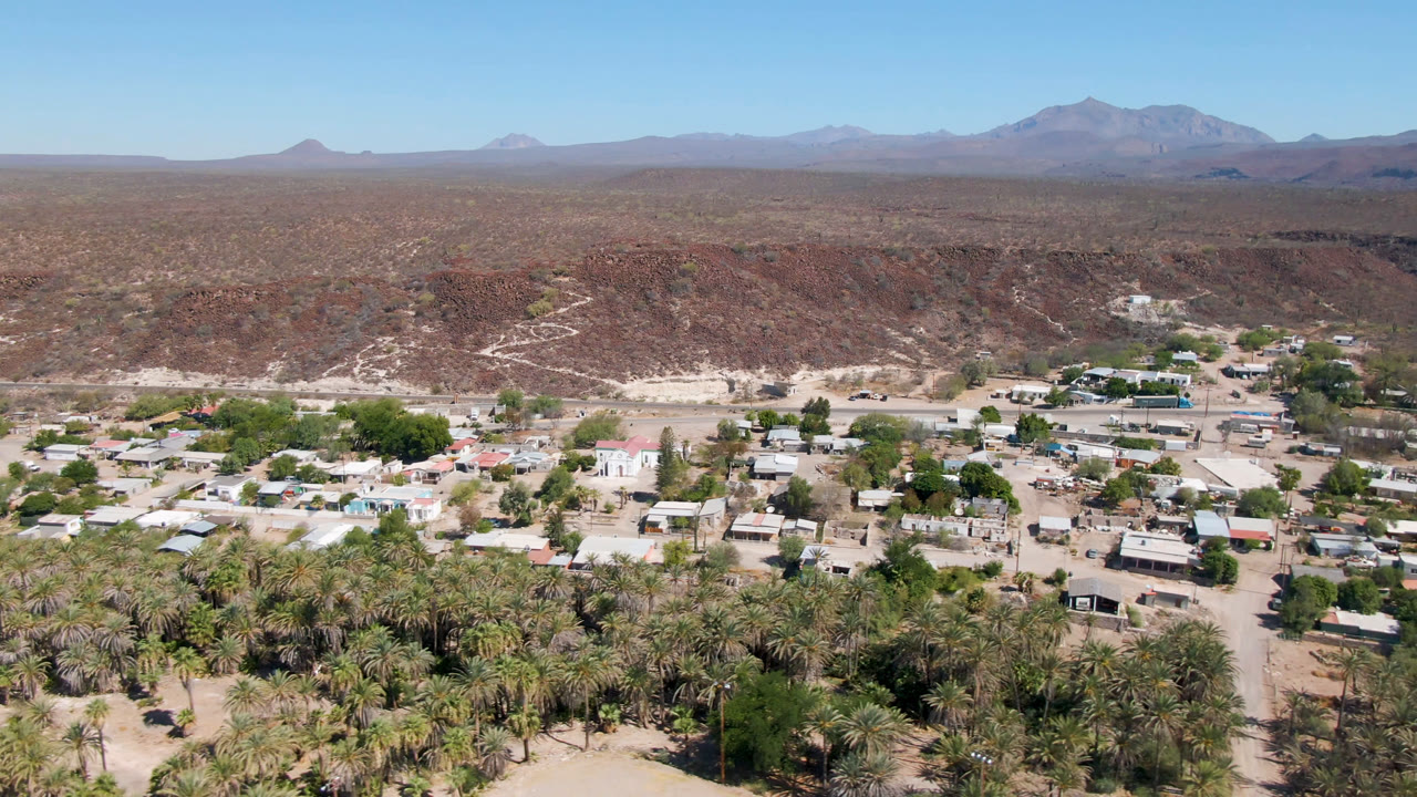 Second aerial frame over San Ignacio showing more of the central settlement, pale dirt roads, palm clusters, red-brown ridges, distant mountains, and no visible people.