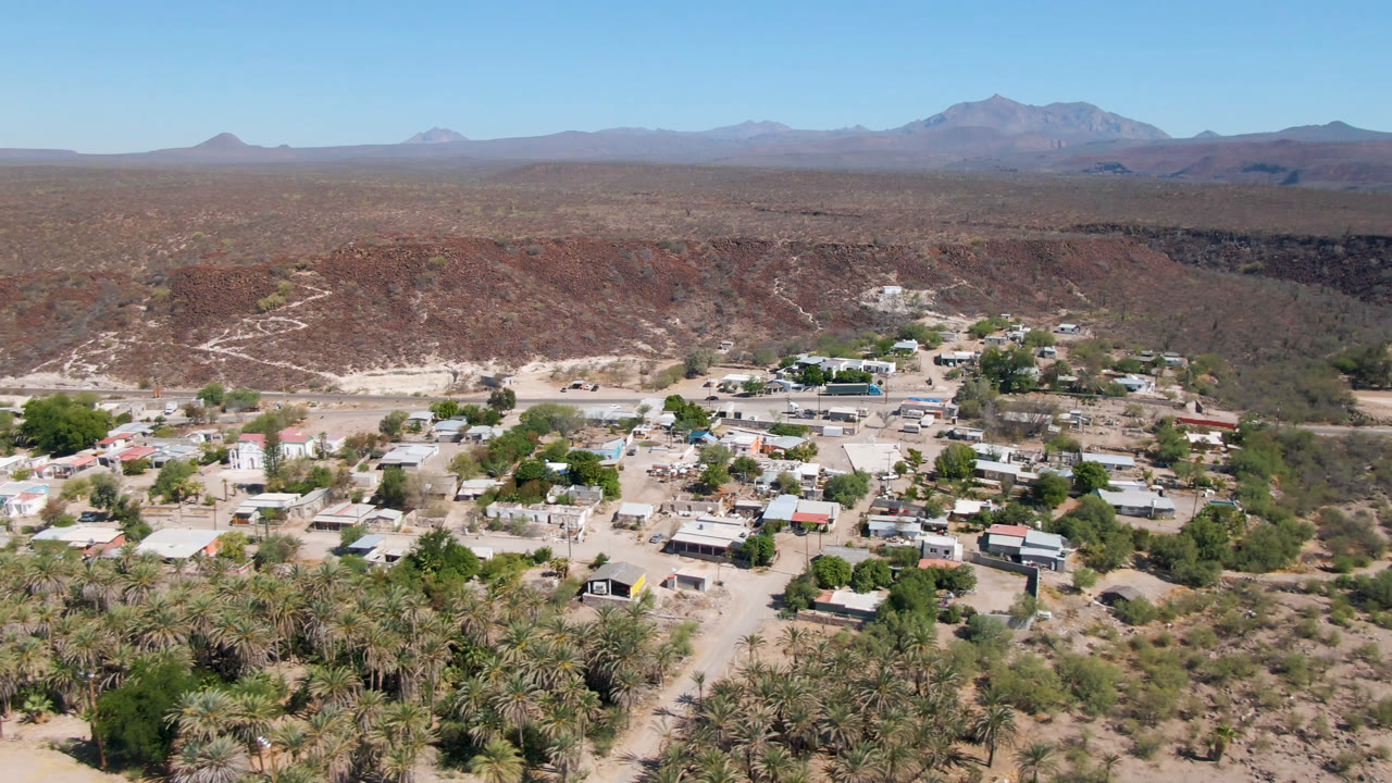 Early aerial view of San Ignacio showing palms in the lower frame, clustered low buildings, pale roads, the desert ridge behind town, distant mountains, and no visible people.