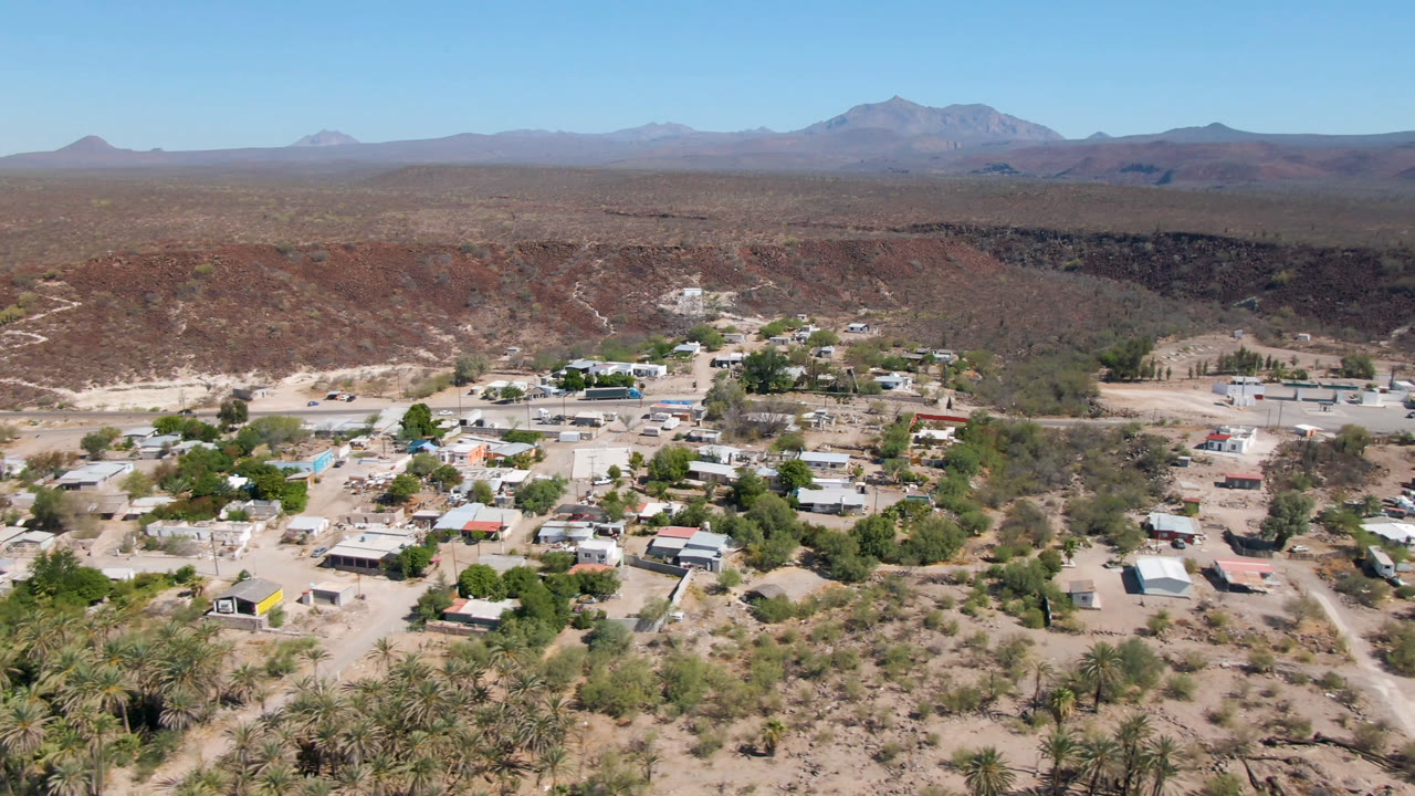 Opening aerial frame over San Ignacio, Baja California Sur, showing low buildings, sandy roads, palm clusters, desert scrub, a dark ravine line, distant mountains, and no visible people.