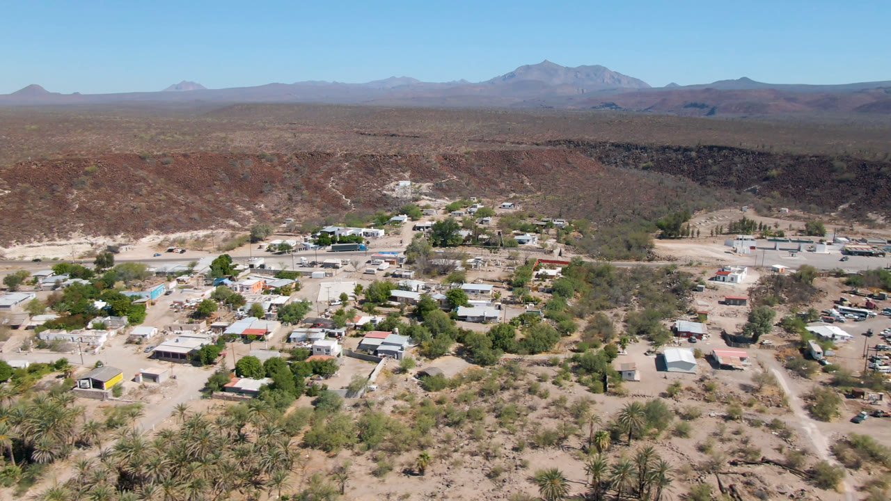 Early aerial view of San Ignacio with scattered roofs and vehicles in the lower frame, green trees around the settlement, dry hills beyond, mountains on the horizon, and no visible people.