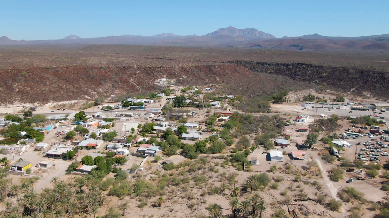 Wide aerial of a small desert settlement with palms, roads, scrub, ravine, and mountains.