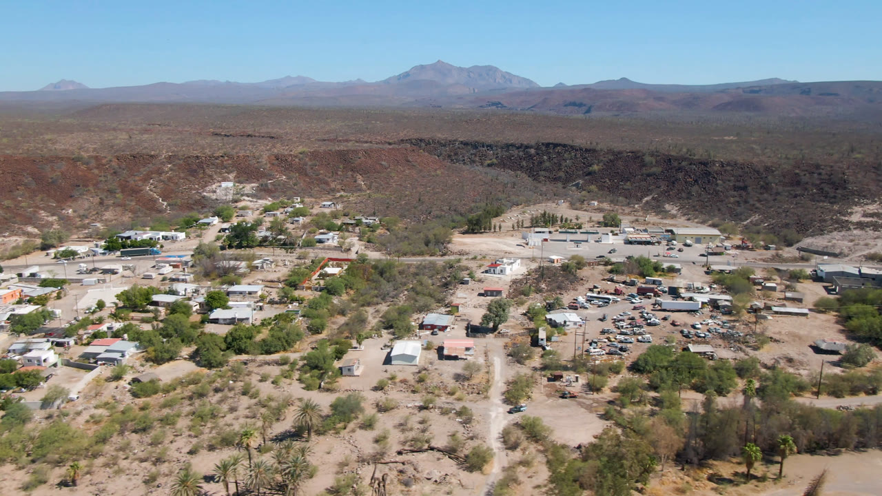 Second aerial frame over San Ignacio with the settlement centered lower in the view, a pale road network, dry scrubland, a shadowed canyon or river corridor, mountains, blue sky, and no visible people.