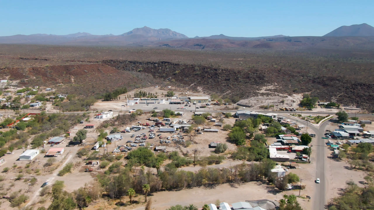 Aerial frame over San Ignacio with low town buildings and parked vehicles below, a road running through the right side, a dark desert ridge behind, and no visible people.