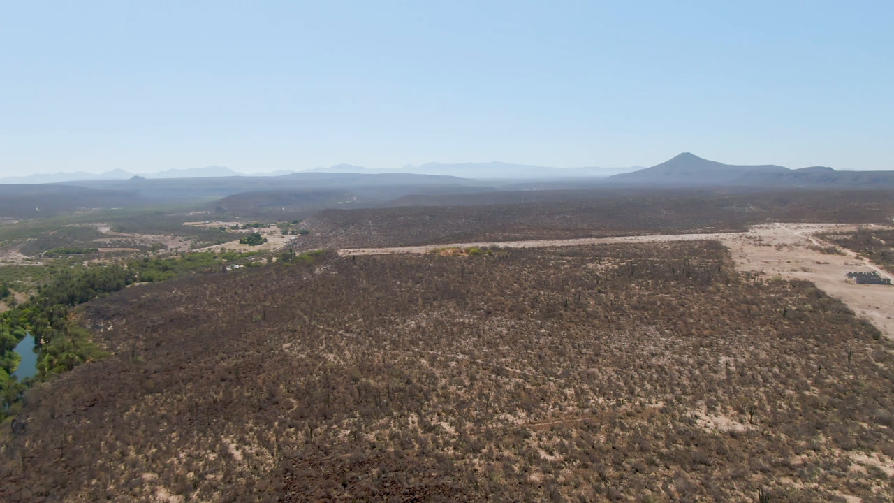 Second aerial frame over San Ignacio, Baja California Sur, with the dirt road more centered, green vegetation along the wash at left, broad brown scrubland, mountain ridges, and no visible people.