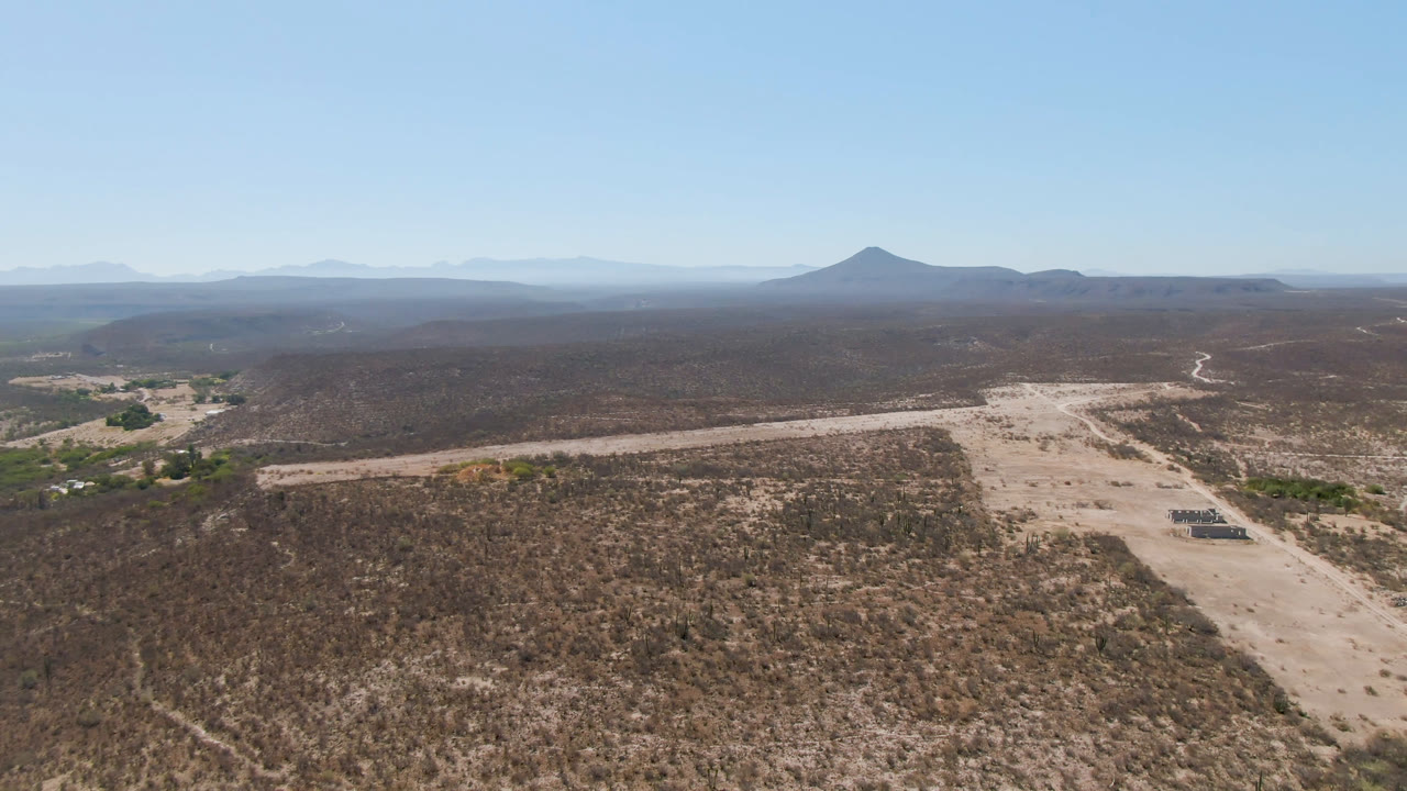 Wide aerial frame in San Ignacio, Baja California Sur, showing a light dirt track across brown desert terrain, sparse green vegetation, hazy mountains, and no visible people.