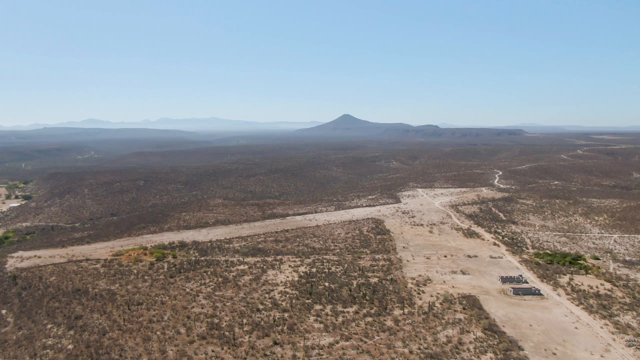 Opening aerial frame over San Ignacio, Baja California Sur, showing a pale dirt road through desert scrub, branching dry washes, distant low mountains, a dark crater-like landform on the horizon, and no visible people.