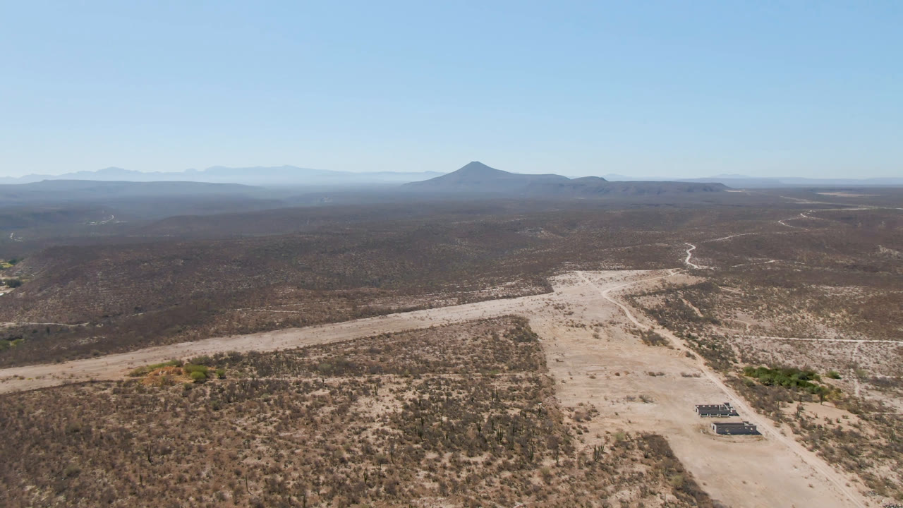 One second into the clip, the San Ignacio desert aerial frames a sandy road and dry wash in the lower half, sparse brown vegetation across the plain, hazy mountains, the distant crater-like formation, and no visible people.