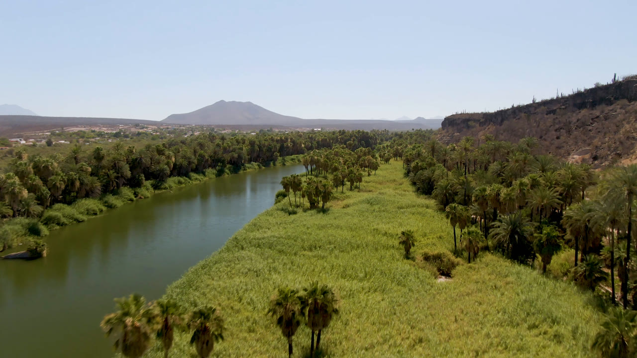 Wide aerial frame in San Ignacio, Baja California Sur, showing the river running through green vegetation with a rocky ridge to the right and distant mountains under clear sky, with no visible people.