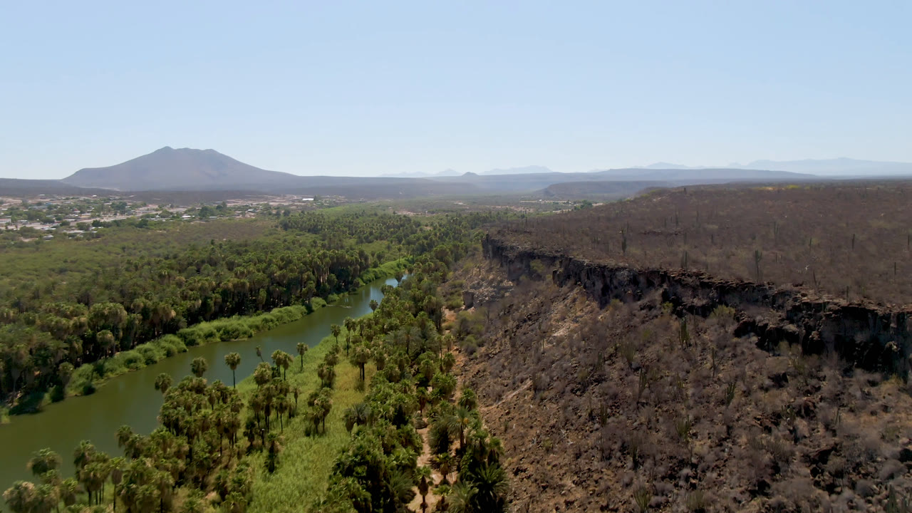 Aerial view over San Ignacio, Baja California Sur, with a green river curving through trees beside dark rocky terrain, distant mountains, clear sky, and no visible people.