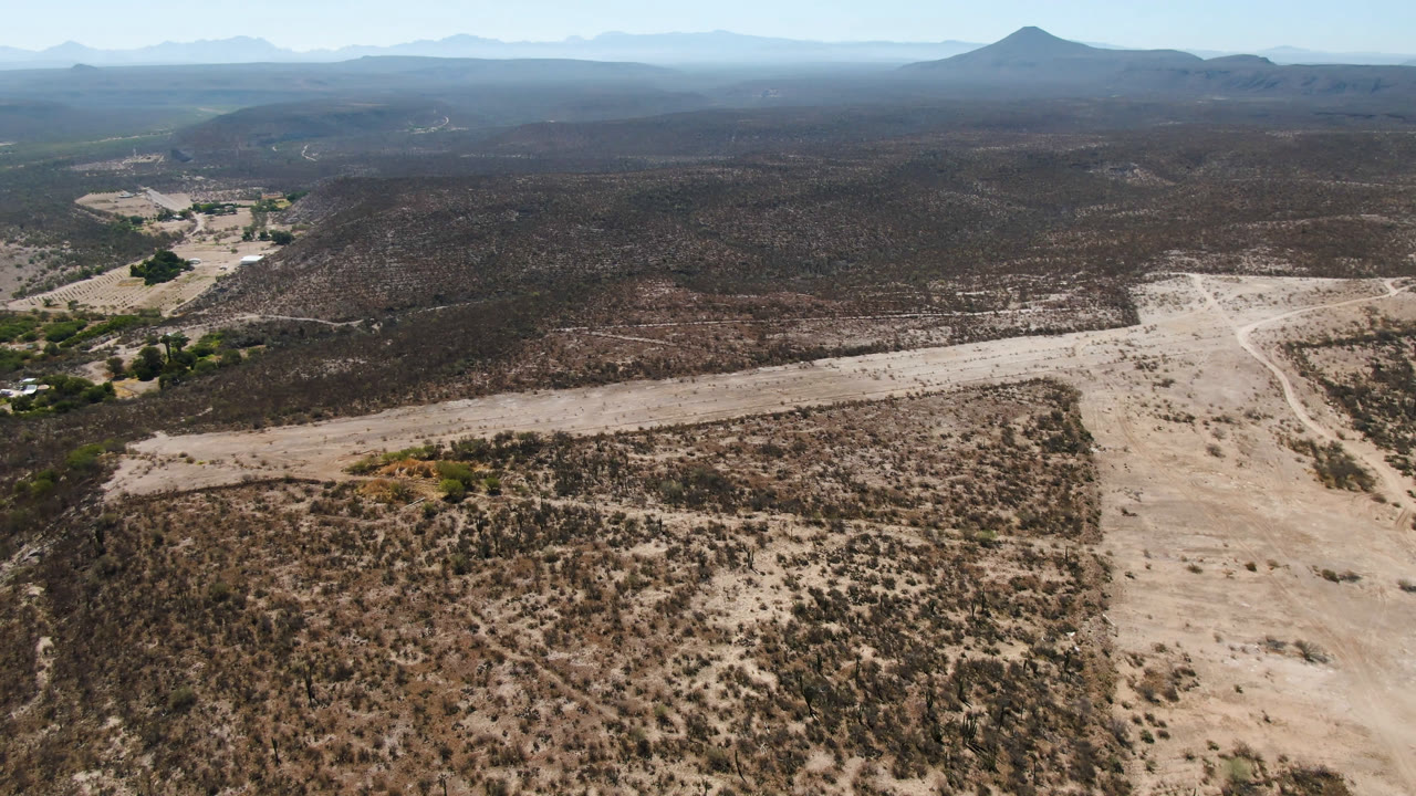 High aerial frame over San Ignacio, Baja California Sur, showing pale dirt roads through dry scrubland, a low hill, distant blue mountains, and no visible people.