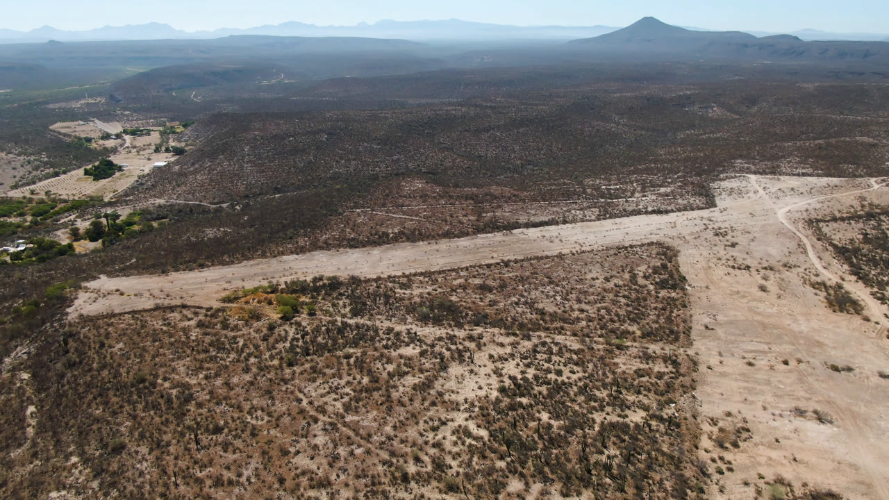 A wide aerial view in San Ignacio, Baja California Sur, frames a pale road bend across the desert floor, scrub-covered rises, distant mountains, and no visible people.
