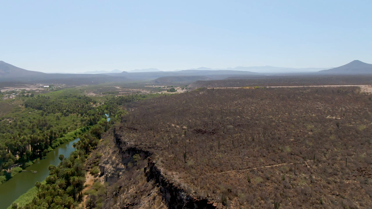 Wide aerial frame over San Ignacio, Baja California Sur, showing a green river and palm corridor at left, a dark dry plateau across the center, distant mountains, clear blue sky, and no visible people.