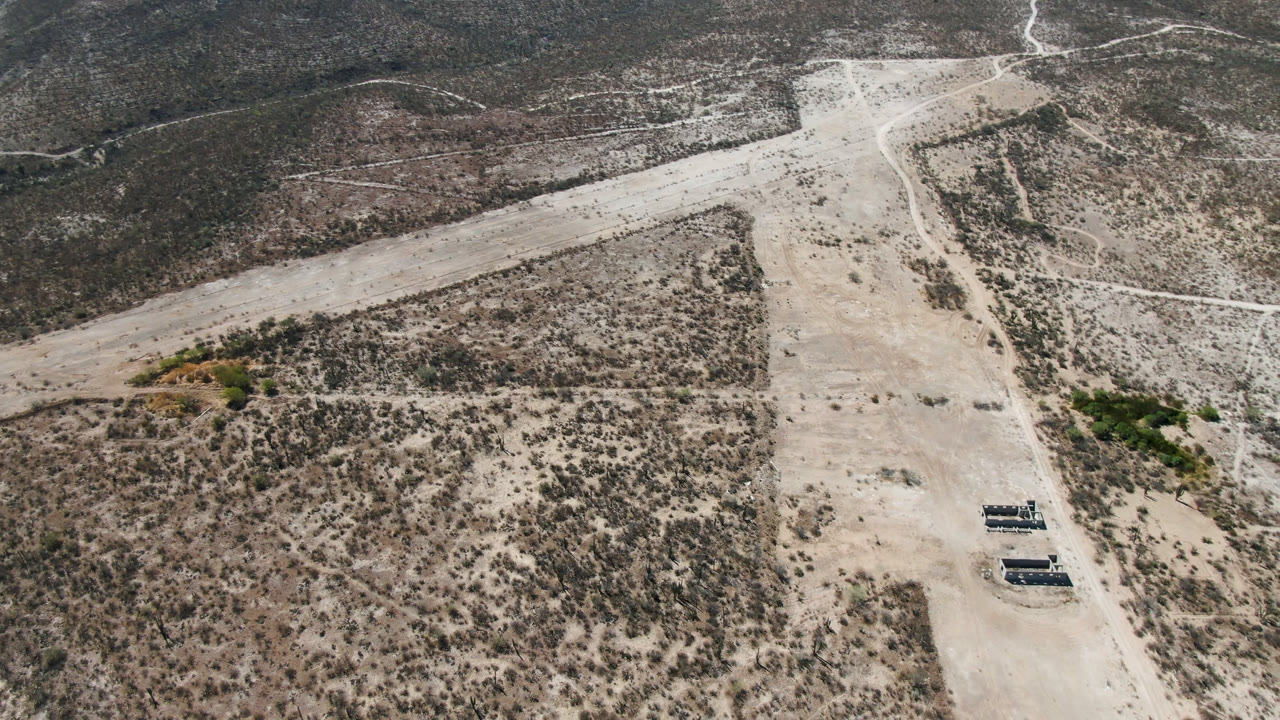 Drone-height view in San Ignacio with intersecting pale dirt tracks, scattered brush, a small green patch, distant hills, and no visible people.