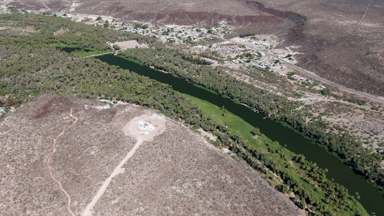 Aerial view over San Ignacio, Baja California Sur, with a dark green river bordered by trees, dry scrubland in the foreground, distant roads and buildings, and no visible people.