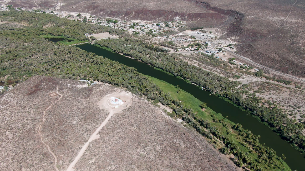 High aerial frame in San Ignacio showing the river bend running diagonally past dense green vegetation, pale desert ground, a dirt access road, distant buildings, and no visible people.