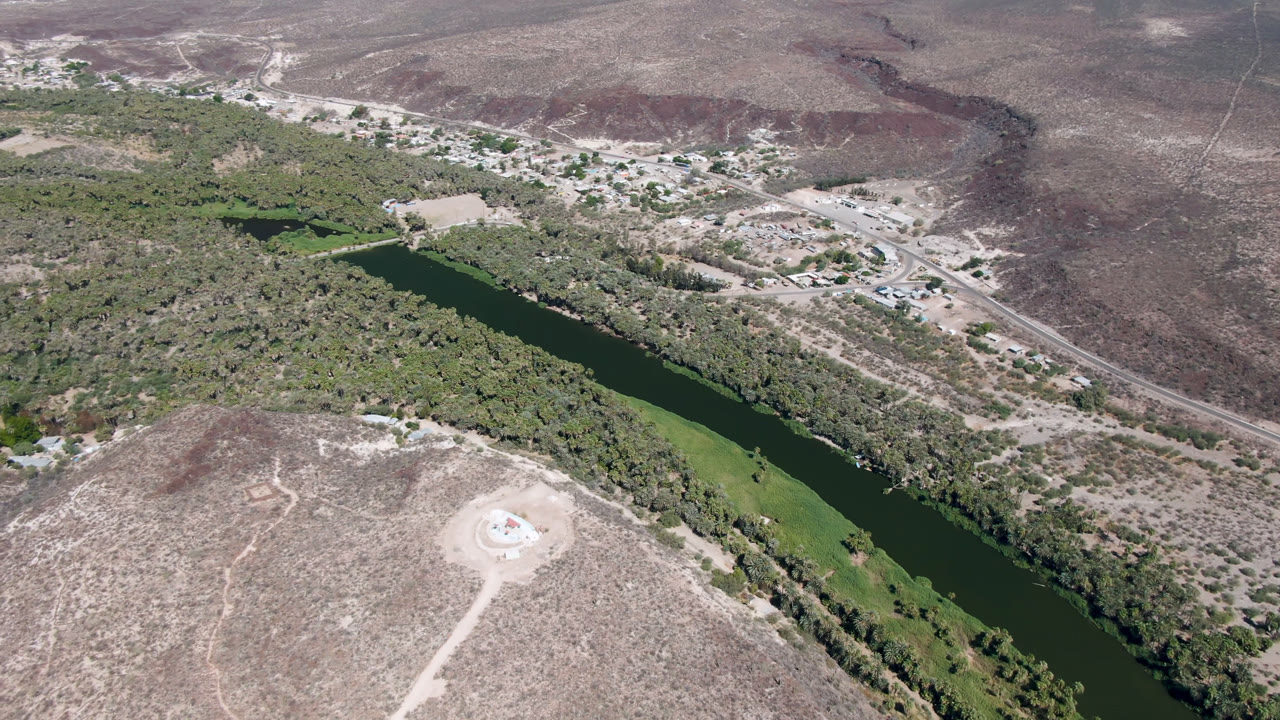 Aerial view in San Ignacio, Baja California Sur, with the river running diagonally past dense trees, a pale hilltop track in the foreground, and no visible people.
