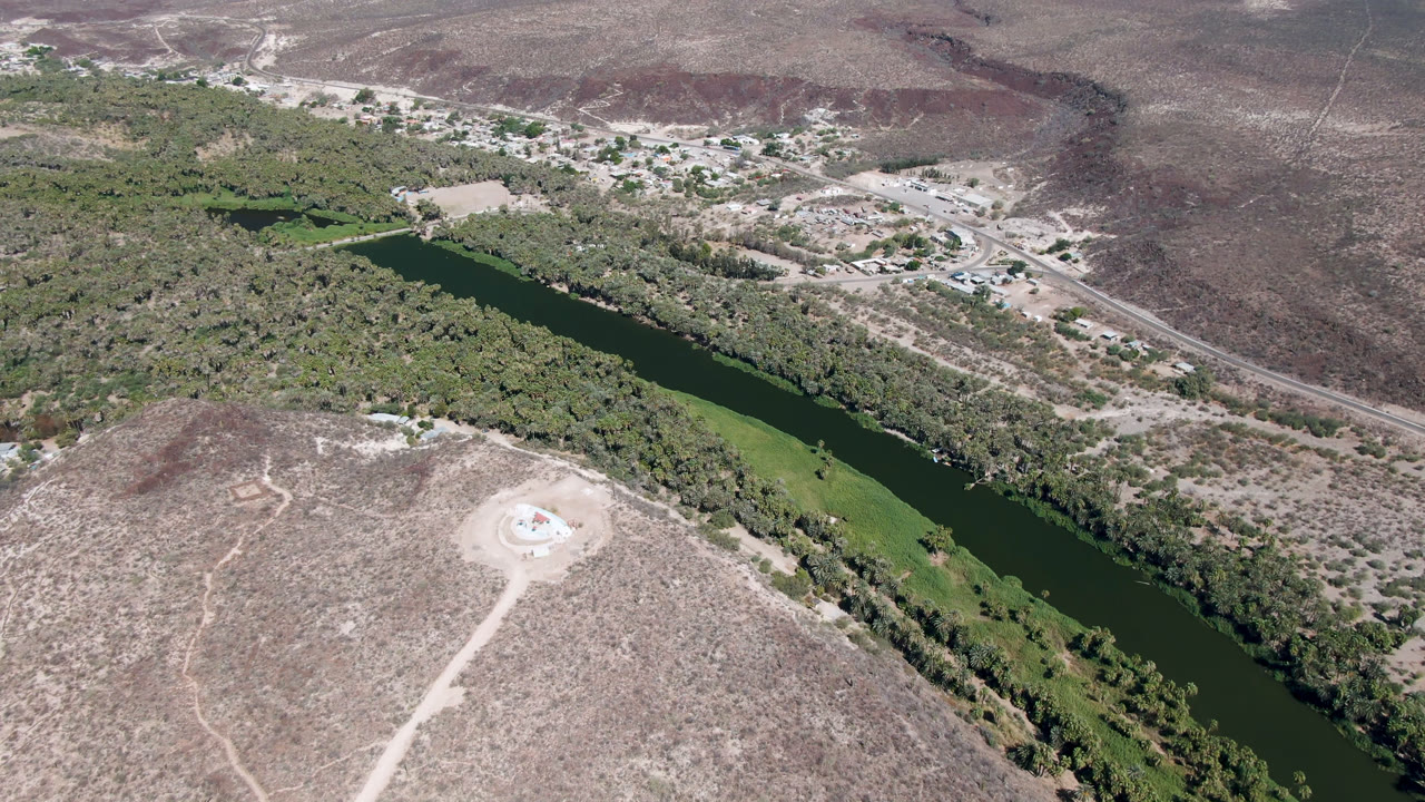 Aerial frame over San Ignacio, Baja California Sur, showing a dark green river running diagonally through dry scrubland, roadside structures in the distance, and no visible people.