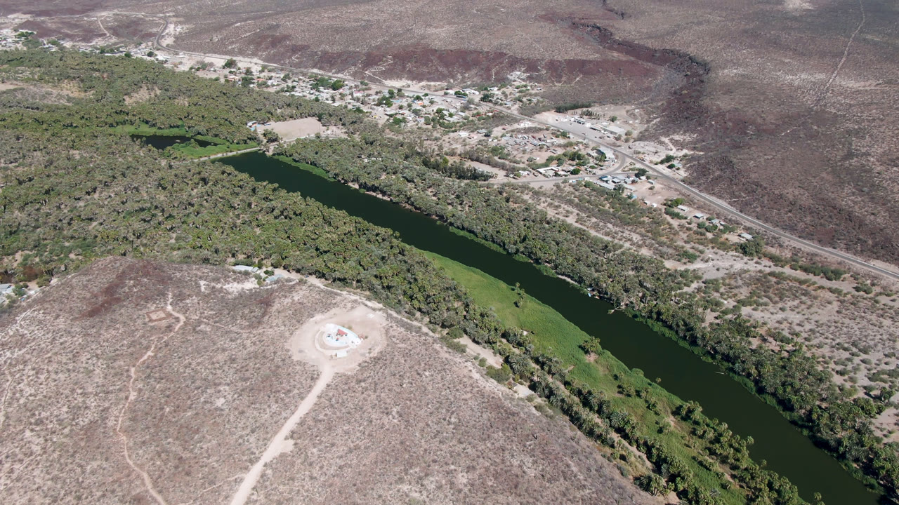 One second into the San Ignacio aerial clip, the river remains centered between green banks, dry hills, pale tracks, and a small developed area beyond the water, with no visible people.
