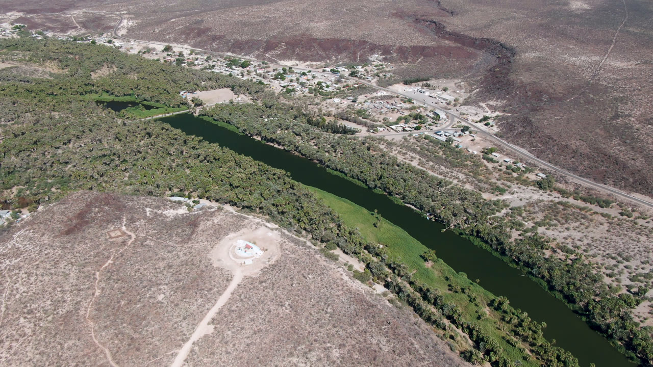 Aerial landscape with a dark river, green banks, dry hills, roads, and a circular feature on a hill.