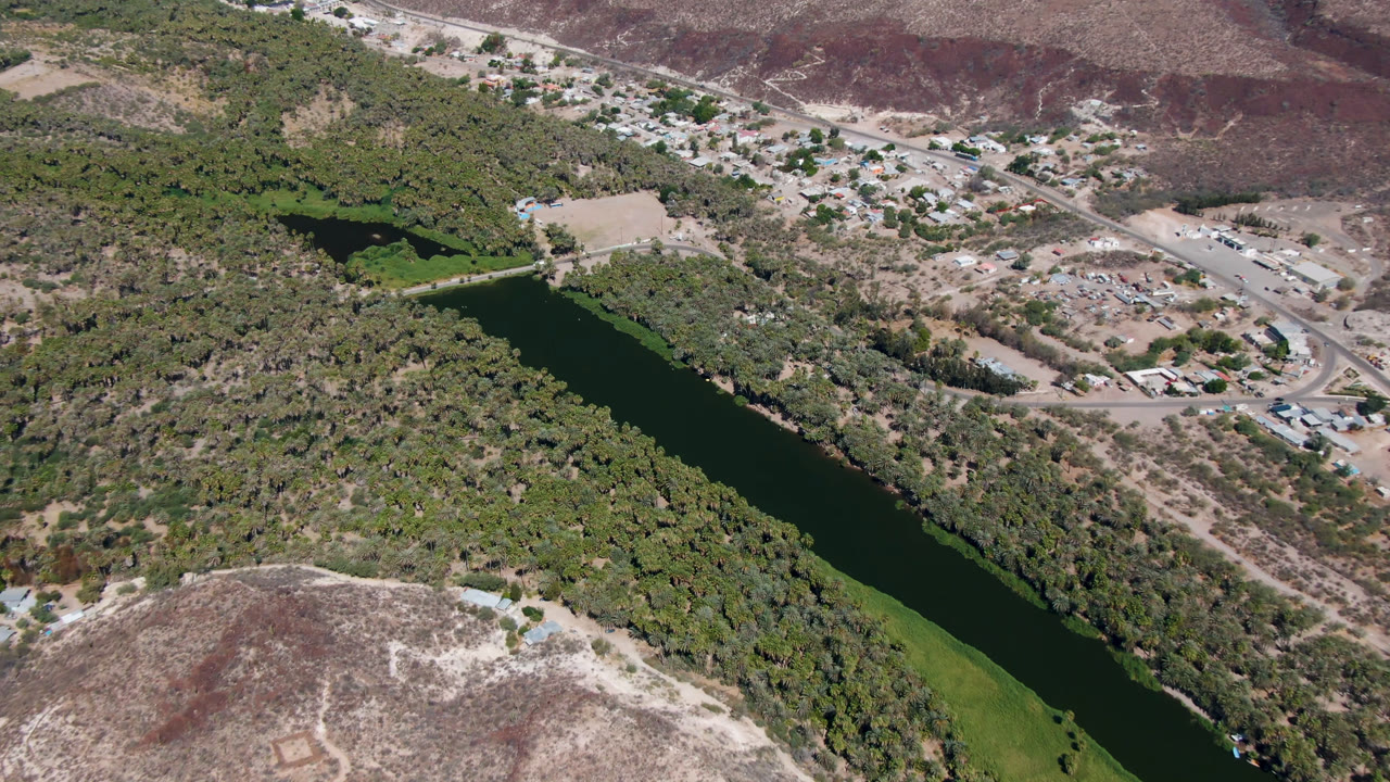 Aerial view in San Ignacio, Baja California Sur with the river running diagonally through thick vegetation, roads and buildings above it, and no visible people.