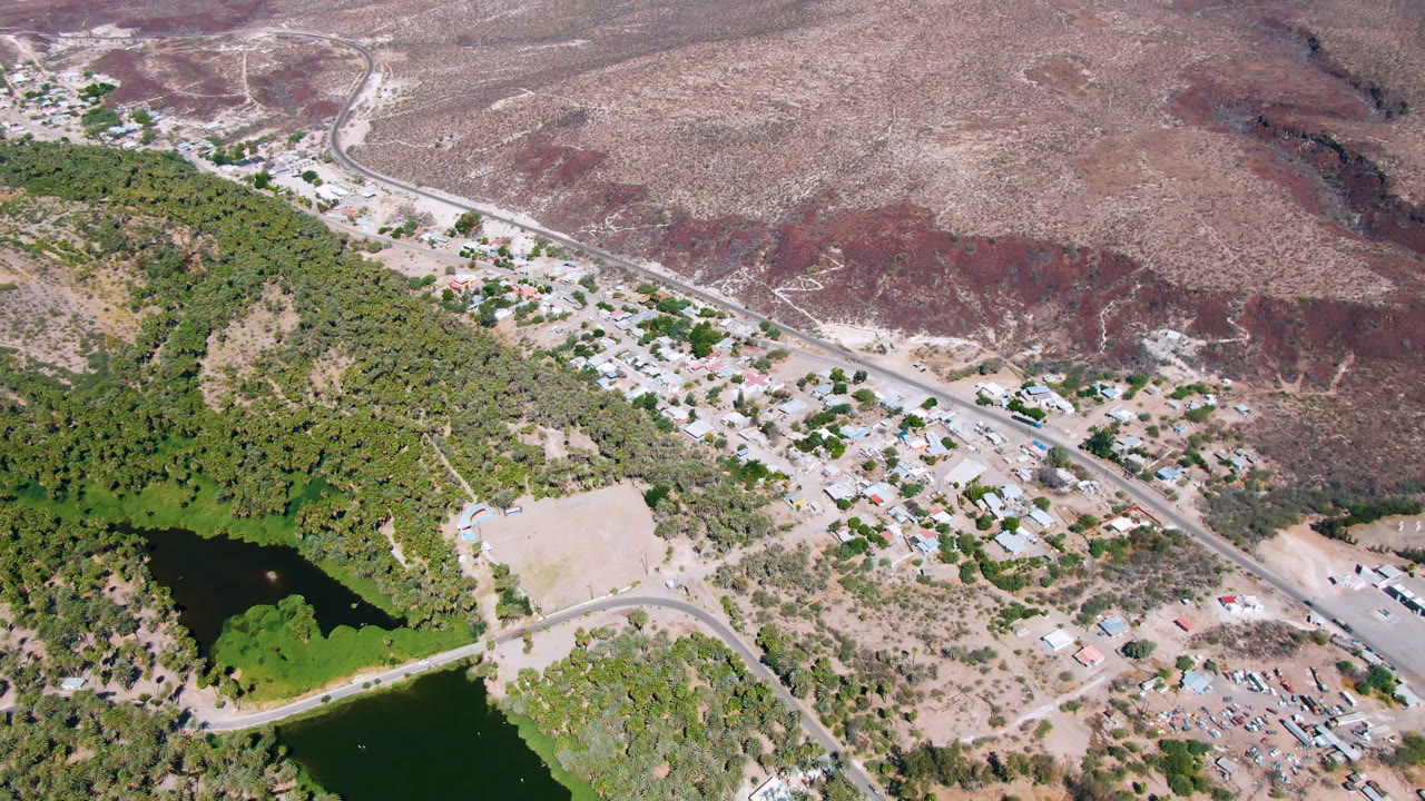 Aerial view of a small town beside water, palms, road, and desert hills.