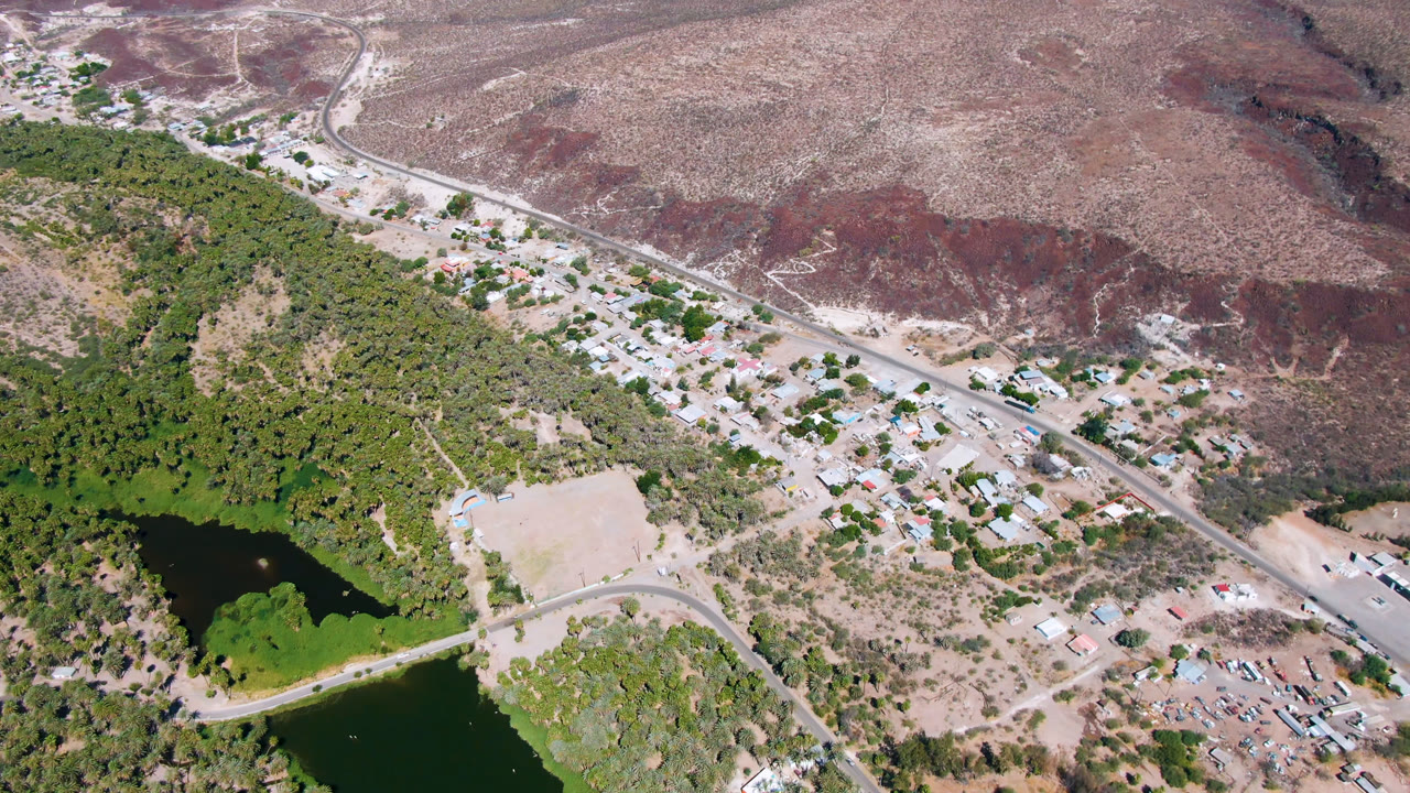 Aerial frame over San Ignacio with the water and palm grove lower in frame, low buildings clustered near the road, pale dirt tracks, reddish hills, and no visible people.