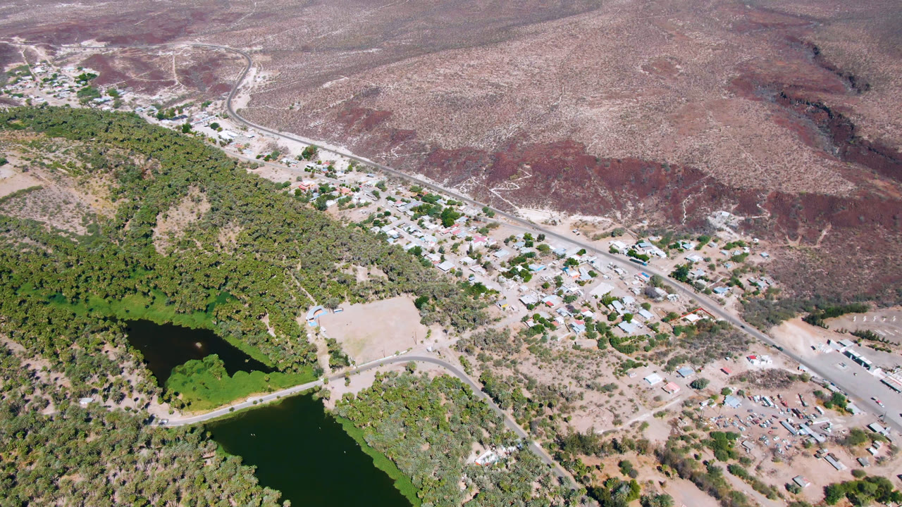 Aerial view over San Ignacio, Baja California Sur with dark green water at lower left, dense vegetation, pale roads, scattered buildings, reddish desert slopes, and no visible people.