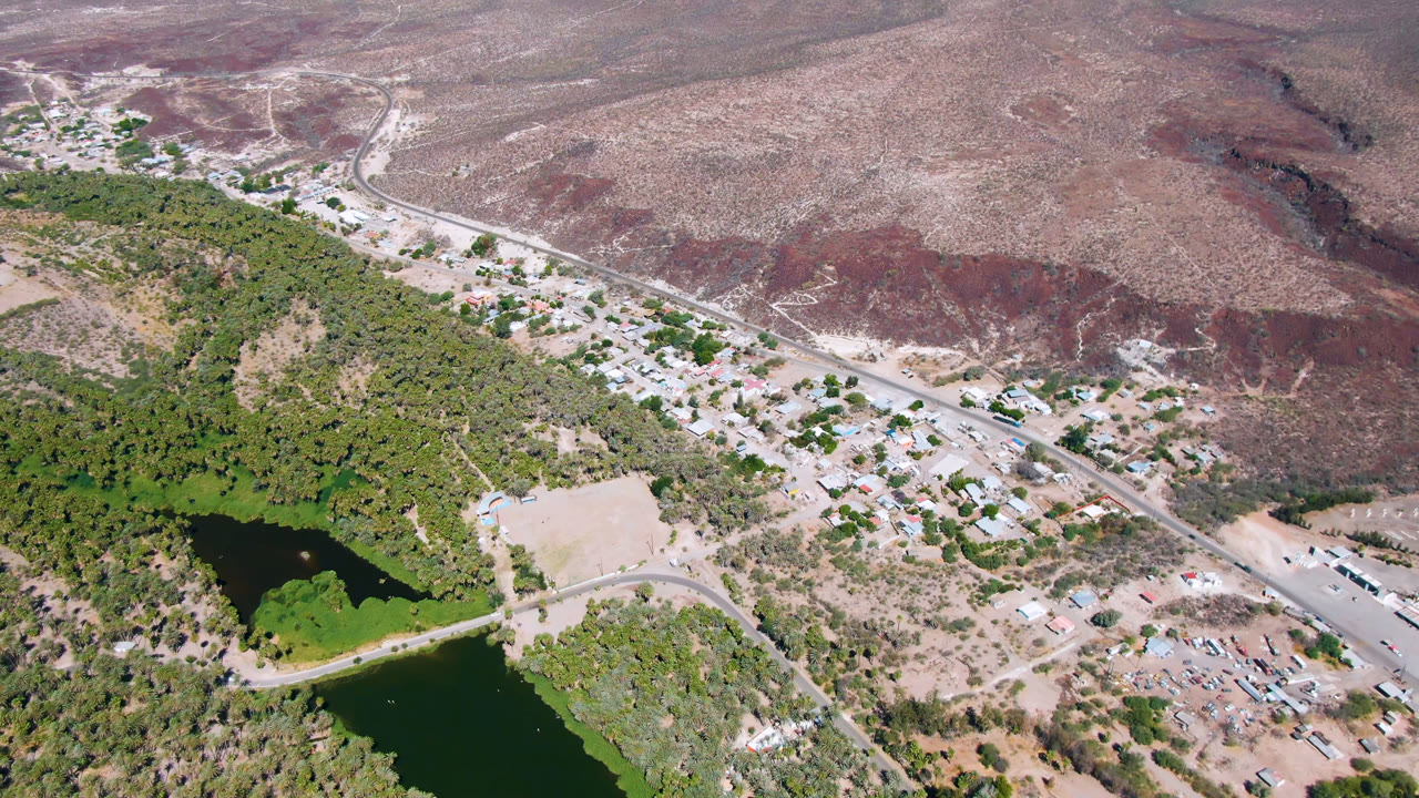 High drone view of San Ignacio with a diagonal road, clustered low buildings, green water and tree cover in the lower left, bare desert terrain beyond, and no visible people.