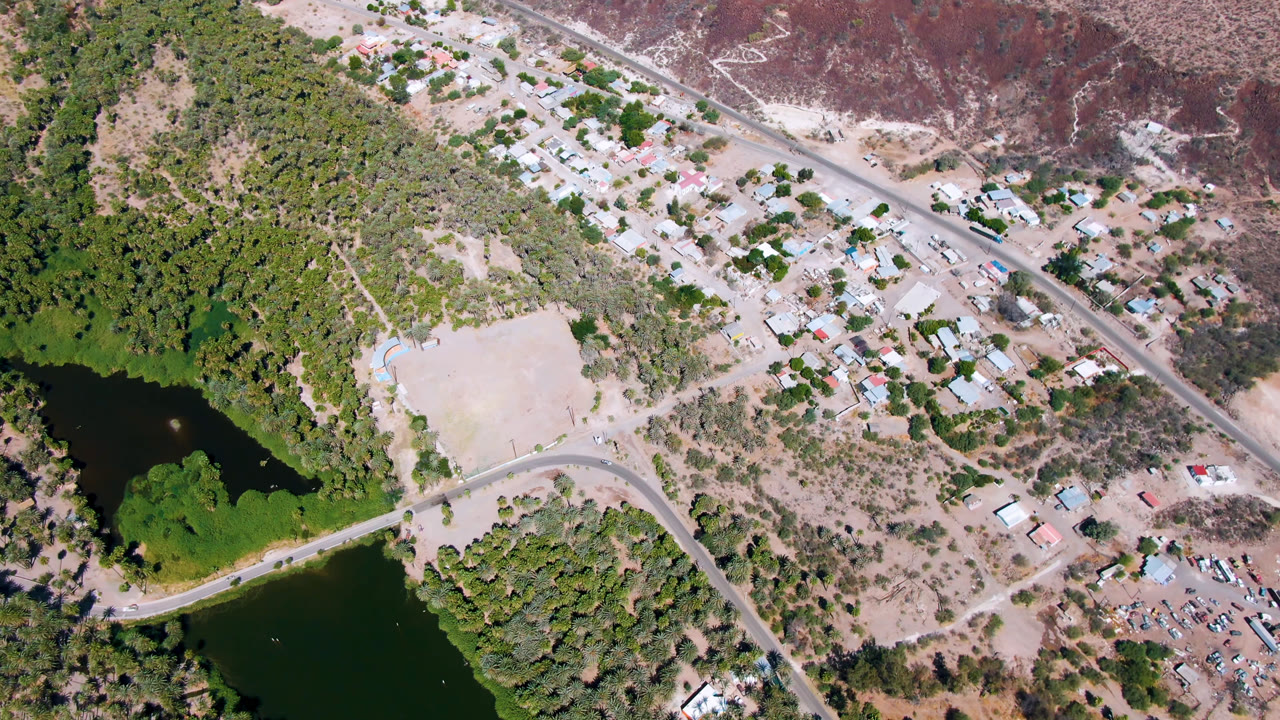 Second overhead frame of San Ignacio, Baja California Sur with the dark water and palms lower left, the road bending across the frame, pale ground near center, buildings beyond it, and no visible people.