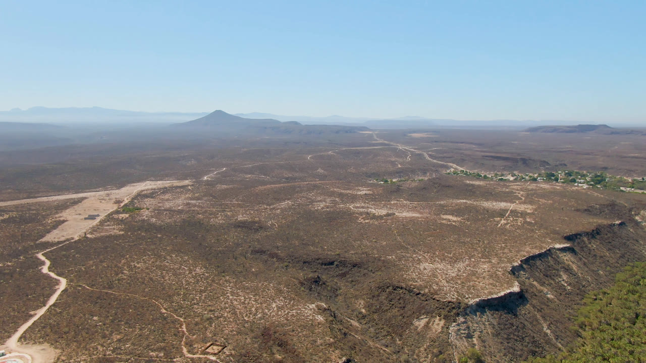 Wide aerial frame over San Ignacio, Baja California Sur, Mexico, showing arid scrubland, pale dirt roads, distant mountains, a darker crater-like rim, a small green area, and no visible people.